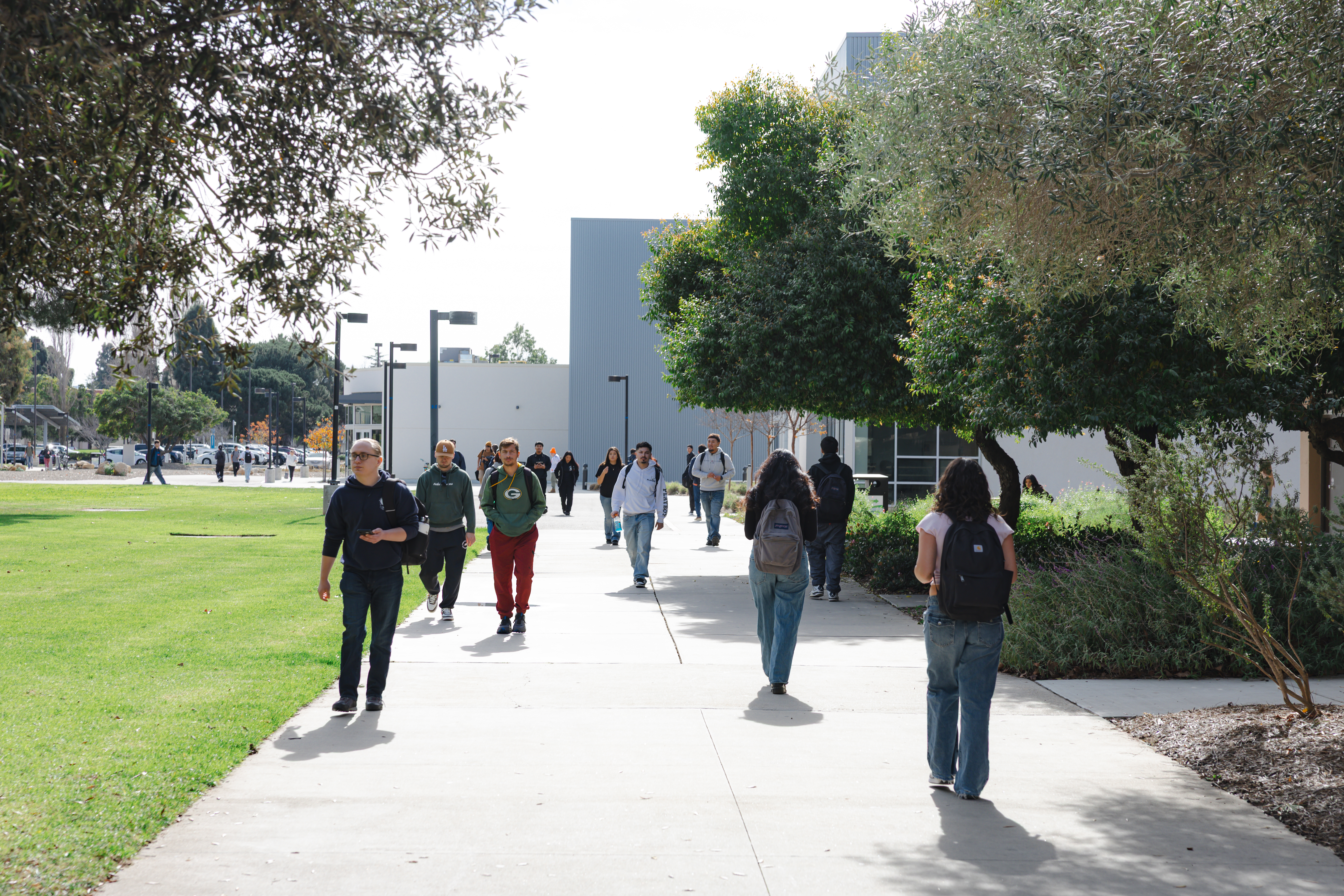 Students walk along a wide pathway at Allan Hancock College, surrounded by trees, lawns and campus buildings on a sunny day.